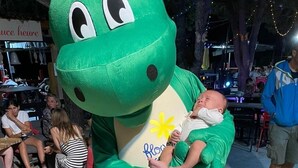 A person in a green dinosaur costume holding a baby.