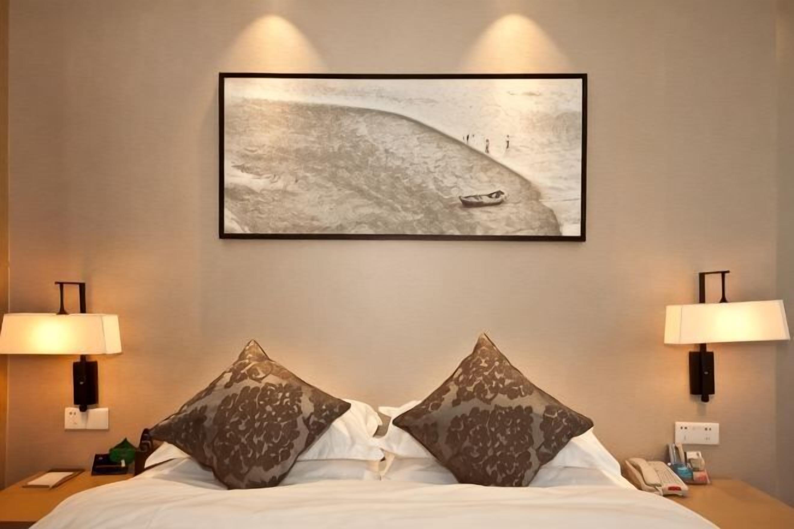 A hotel room with a bed, two lamps, a painting of a boat on water, and a nightstand with a phone.