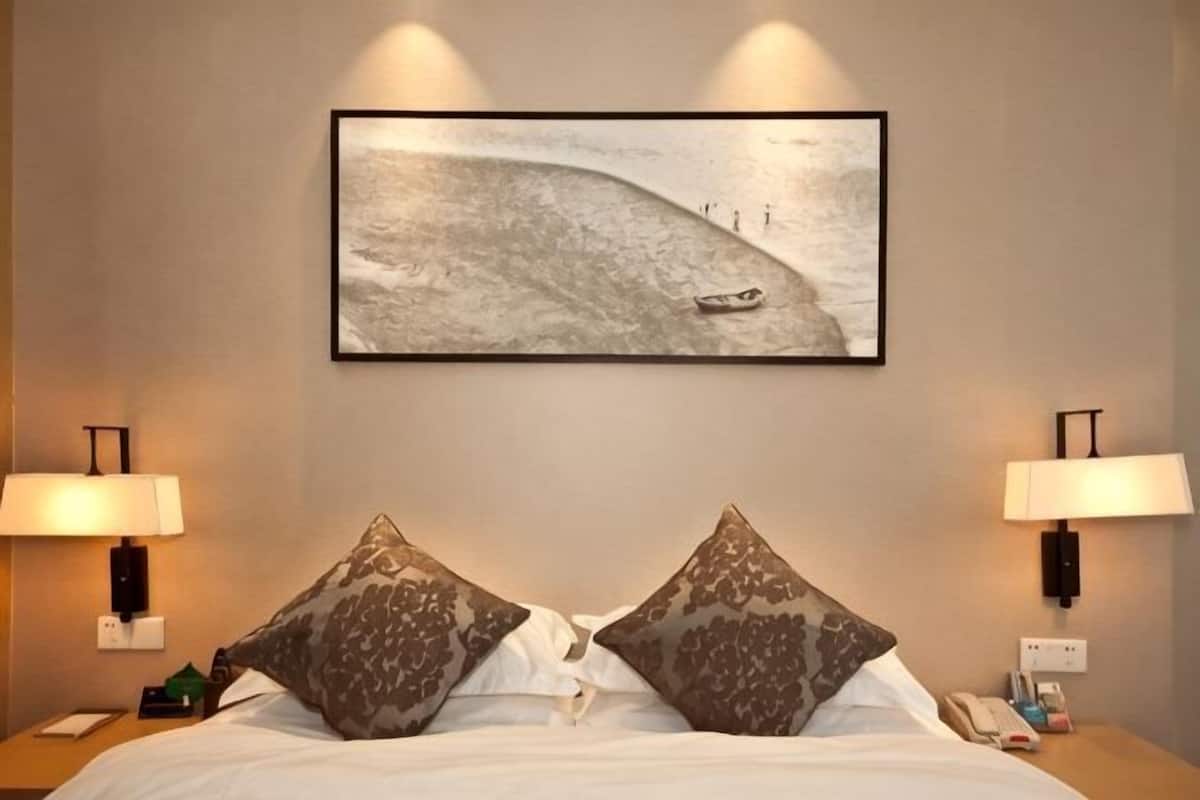Una camera d'albergo con un letto, due lampade, un quadro raffigurante una barca sull'acqua e un comodino con un telefono.