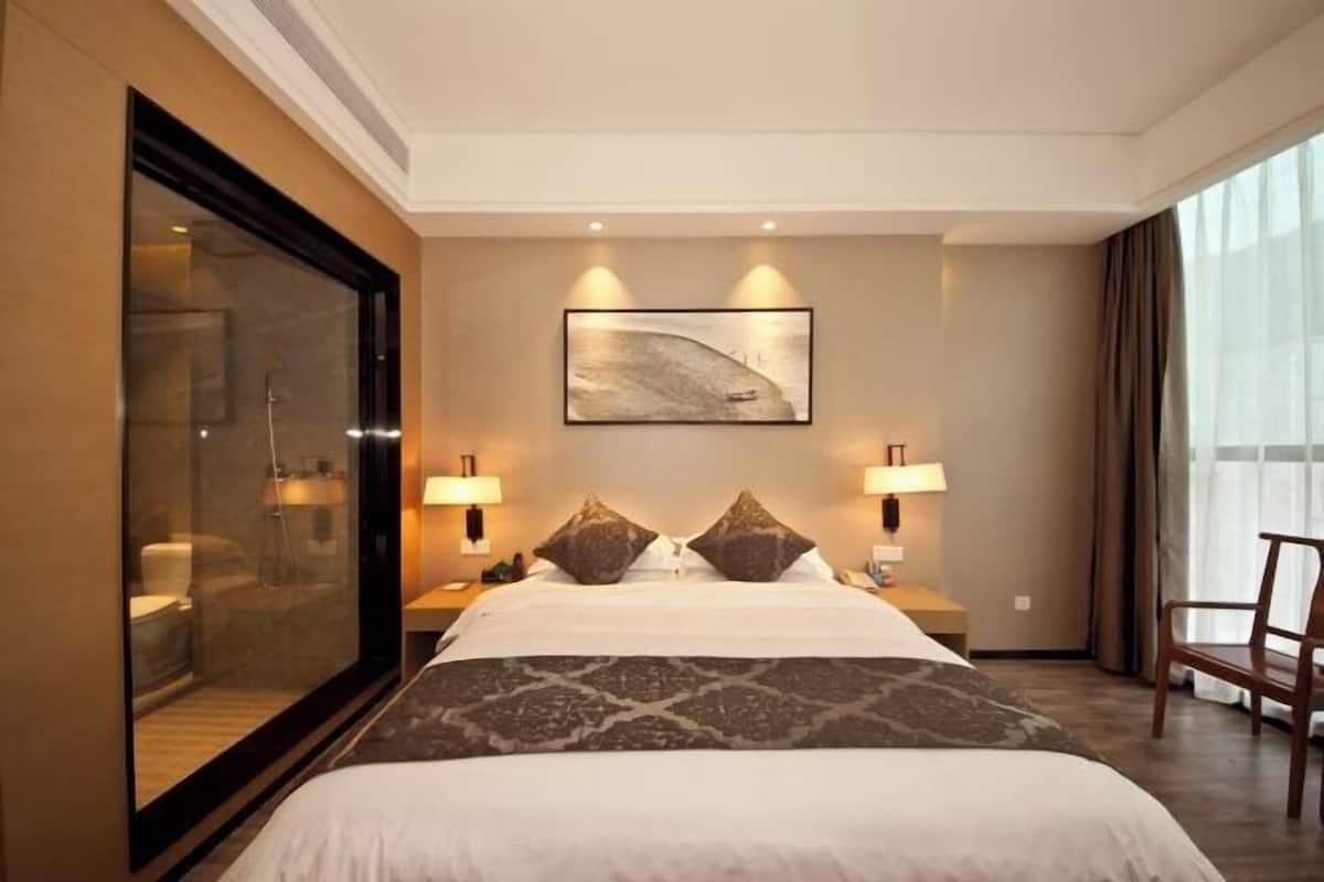 Habitación de hotel con una cama grande, dos lámparas de noche, un amplio ventanal con cortinas y una obra de arte enmarcada en la pared.