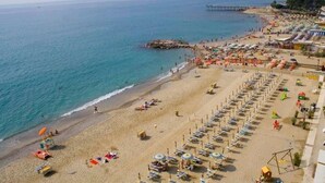 Une plage bordée de rangées de chaises longues et de parasols, une jetée qui s’avance dans la mer et un promontoire rocheux qui émerge de l’eau.