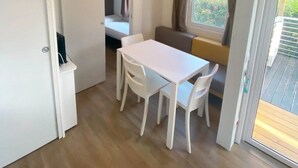 Une pièce de taille modeste, avec une table et des chaises blanches, un banc, une fenêtre avec des rideaux et une porte donnant sur une autre pièce.