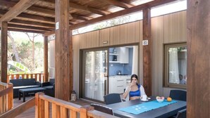 Ein überdachter Außenbereich mit Holzterrasse, Tisch mit Stühlen und Blick auf eine Küche durch eine Glastür.