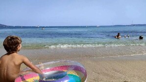 Escena de playa con agua cristalina, un niño sosteniendo un flotador de colores y varias personas nadando a lo lejos.