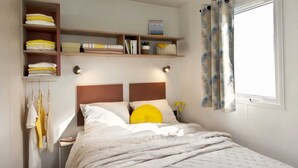 Un dormitorio ordenado con cama, una estantería con toallas dobladas y una ventana con cortinas.
