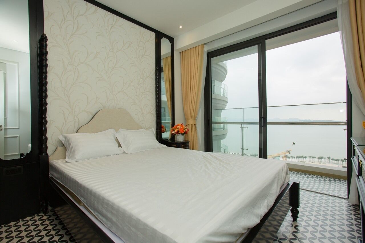 A hotel room with a large bed, a nightstand, a mirror, and a view of the sea through a large window.