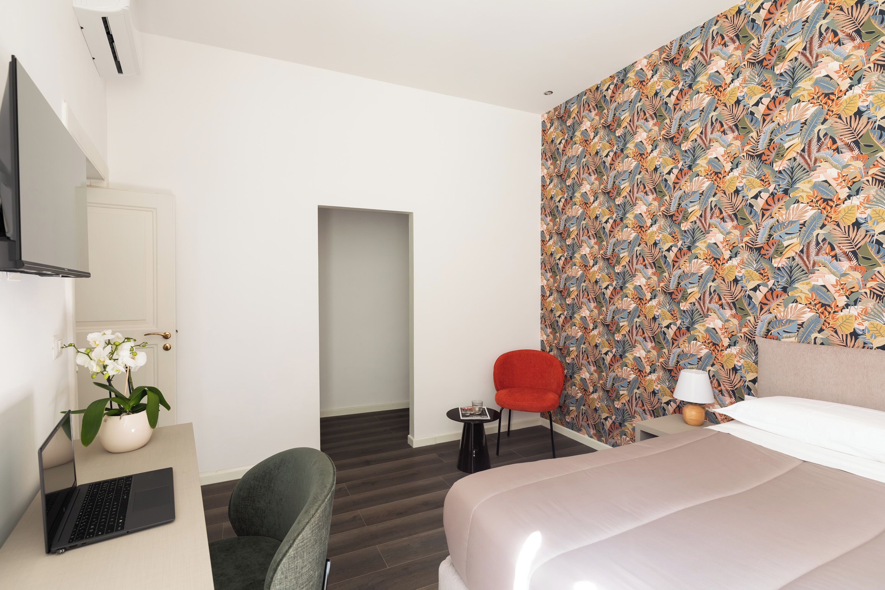 Una habitación de hotel moderna con cama, escritorio con ordenador portátil, una silla roja, una mesita con lámpara y un papel tapiz floral.