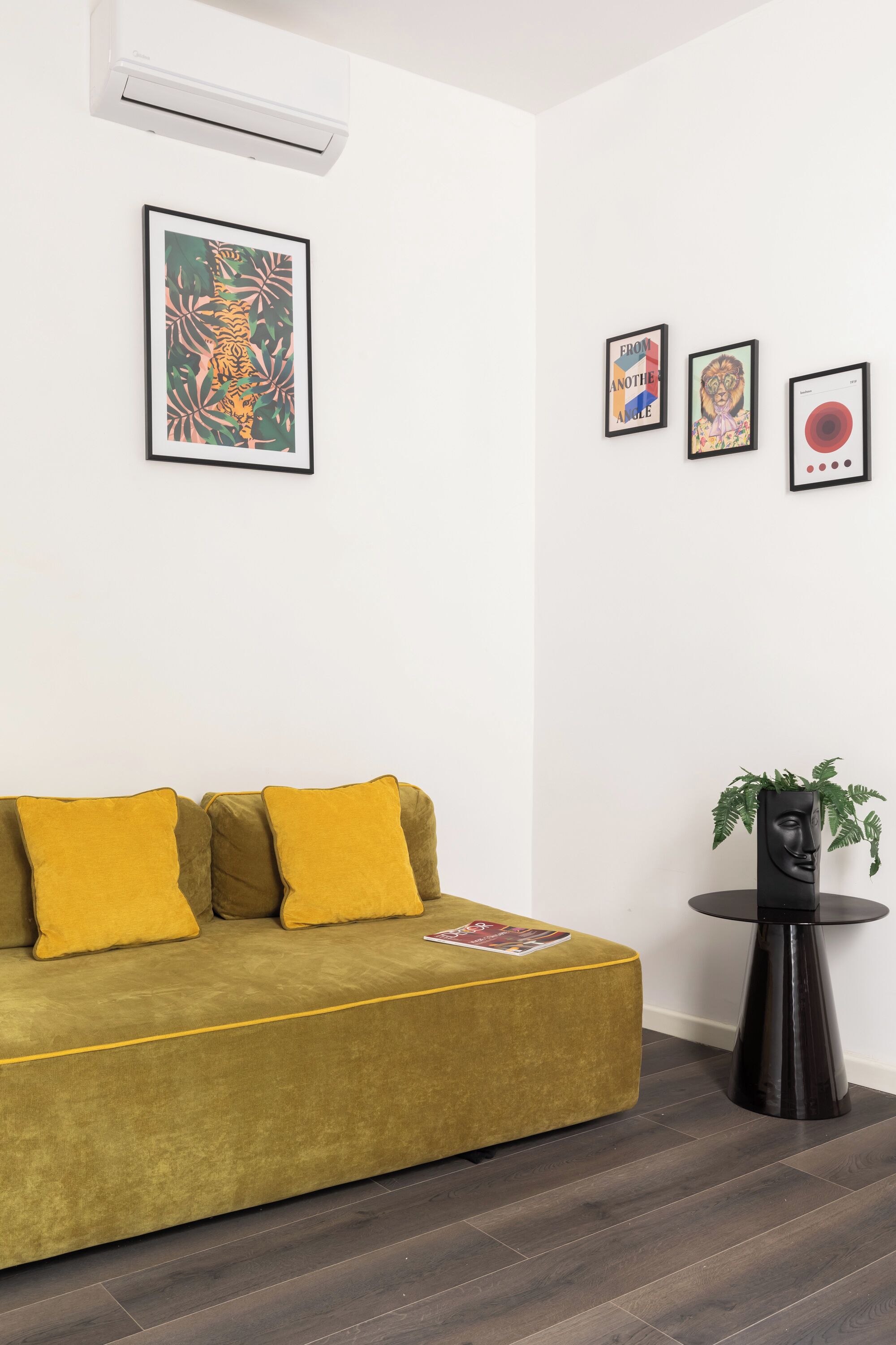 Une pièce avec un canapé vert, une petite table noire sur laquelle se trouve une plante, et trois œuvres d’art encadrées accrochées au mur.