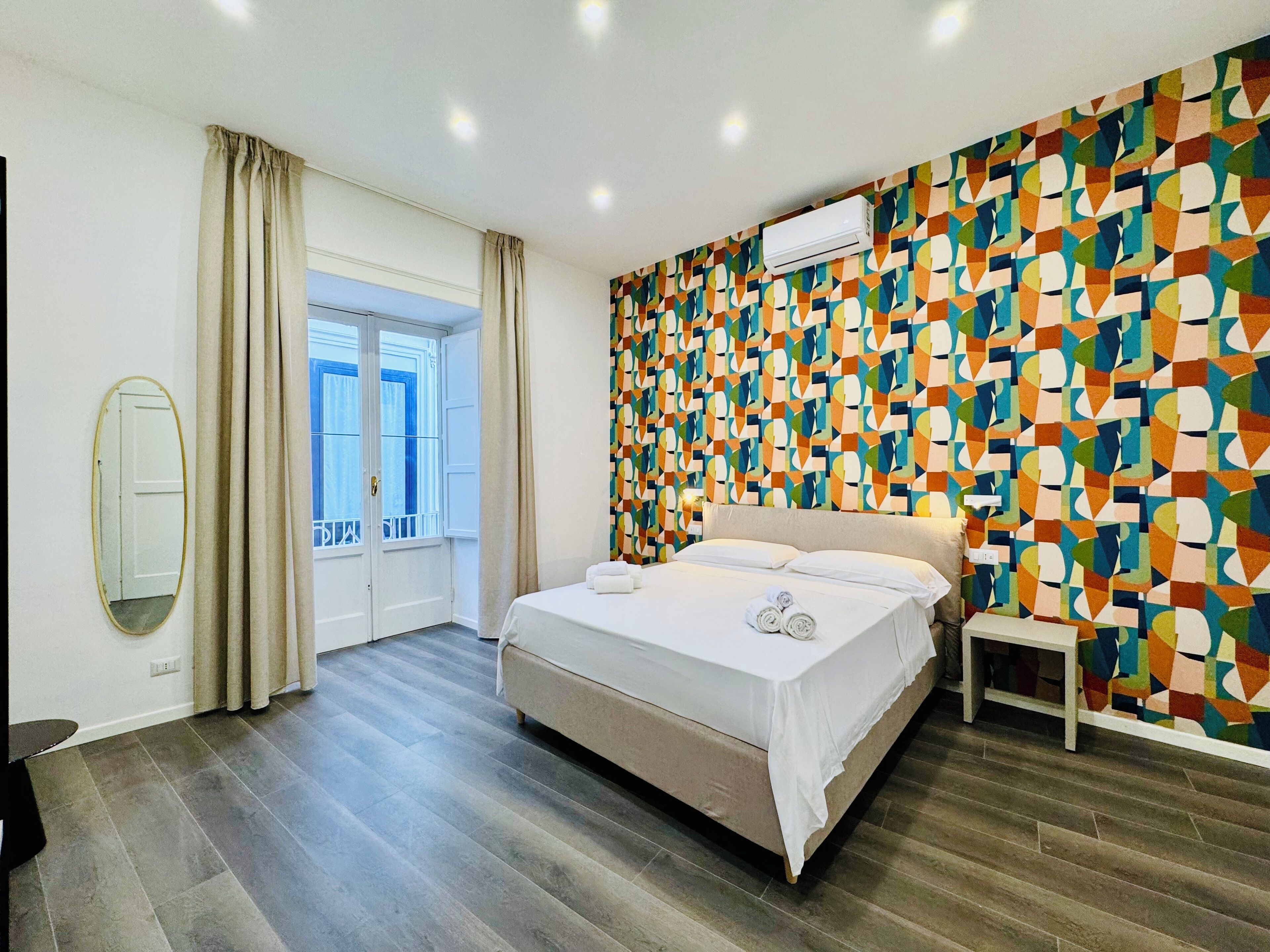 Moderni hotellihuone, jossa on suuri sänky, ikkuna verhoilla, värikäs geometrinen seinäkuvio ja pieni yöpöytä.