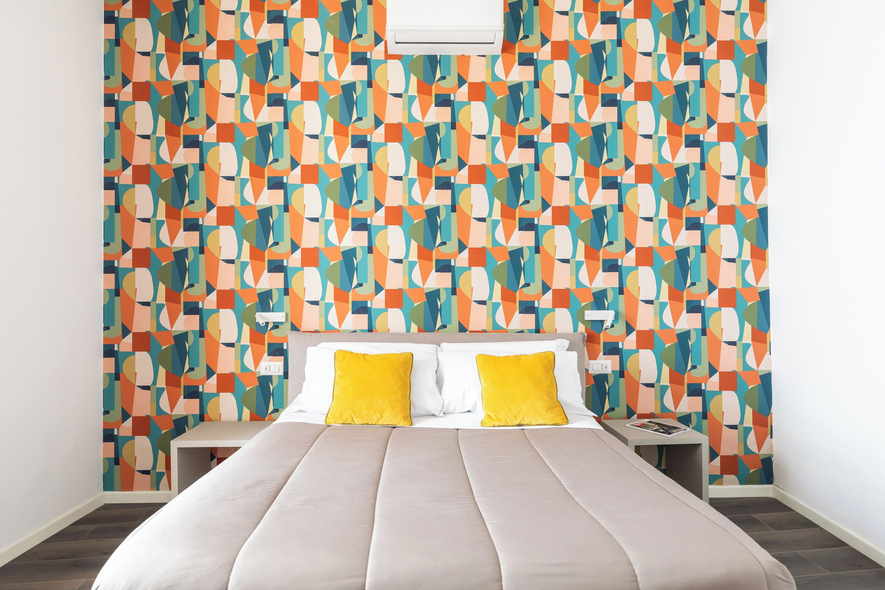 Een hotelkamer met een wand met een geometrisch patroon, een bed met twee kussens en een nachtkastje met een boek.