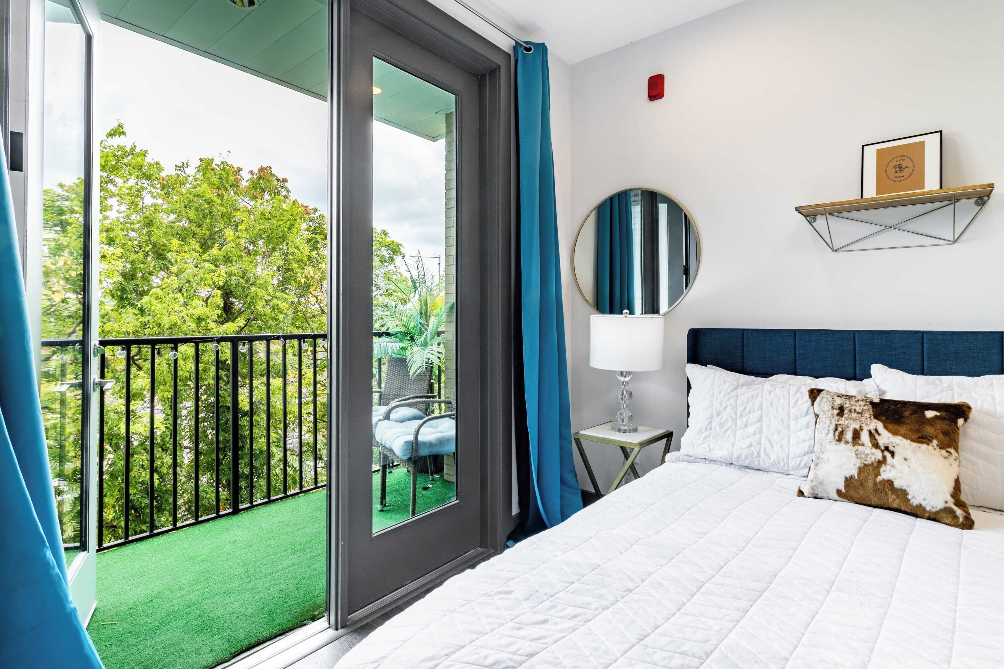 Habitación de hotel con balcón, cama con ropa de cama blanca, espejo redondo y estante montado en la pared con un cuadro enmarcado.