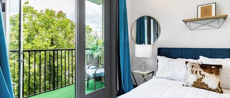 Habitación de hotel con balcón, cama con ropa de cama blanca, espejo redondo y estante montado en la pared con un cuadro enmarcado.