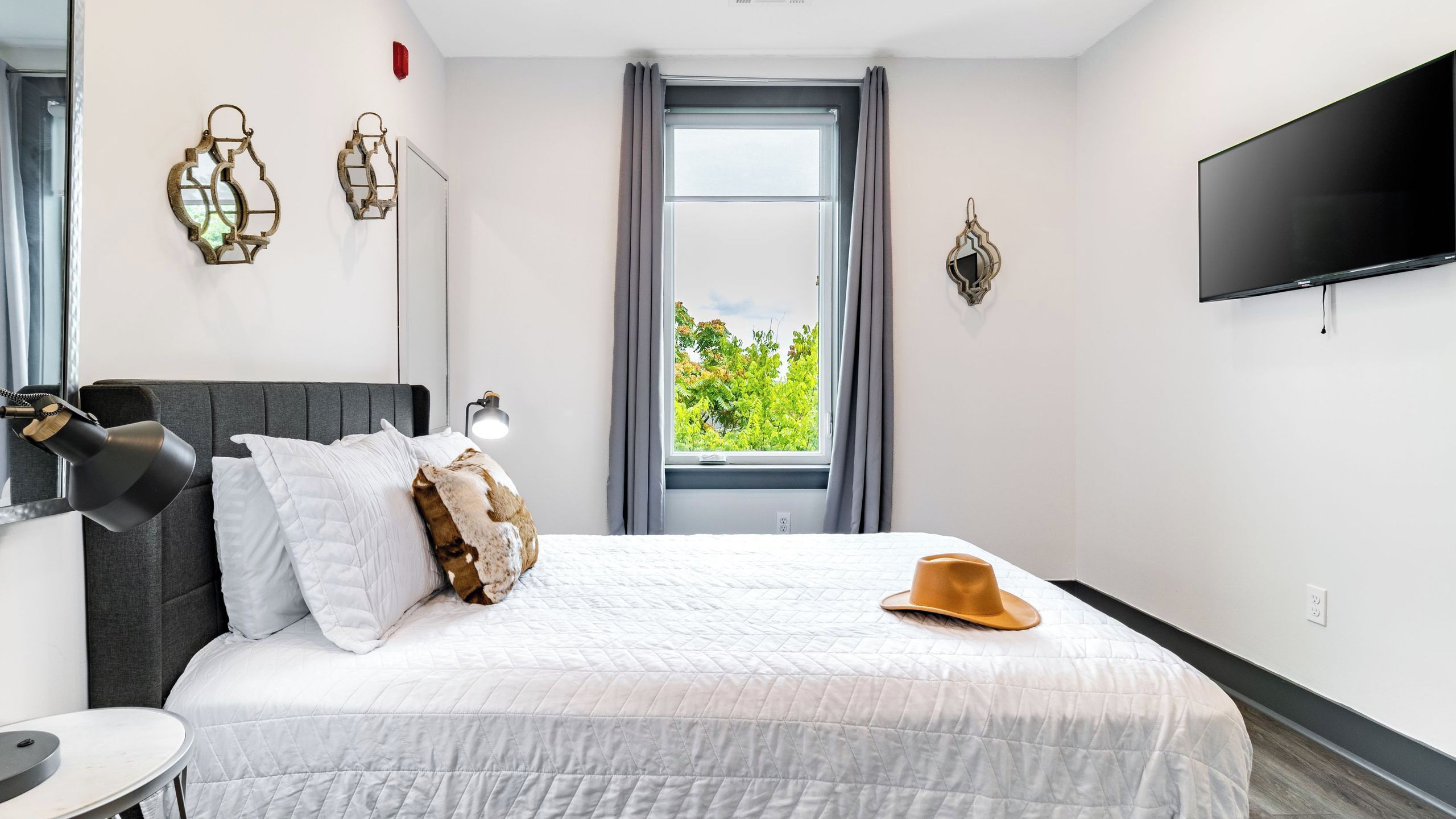 A bedroom with a bed, a bedside lamp, a window with curtains, a wall-mounted TV, and decorative wall sconces.