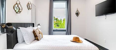 A bedroom with a bed, a bedside lamp, a window with curtains, a wall-mounted TV, and decorative wall sconces.