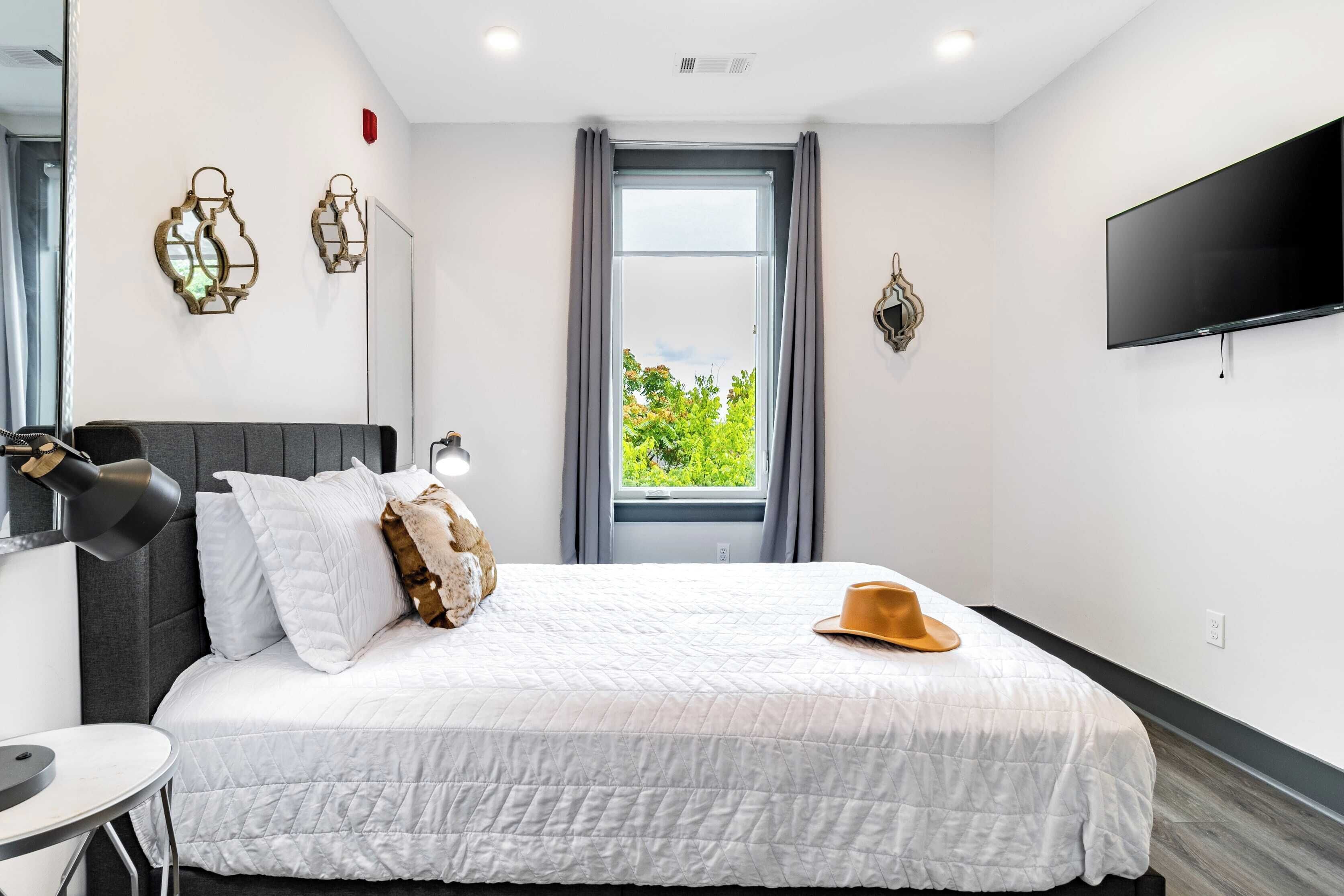 A hotel room with a bed, a desk, a chair, a TV, and a window with a view of greenery.