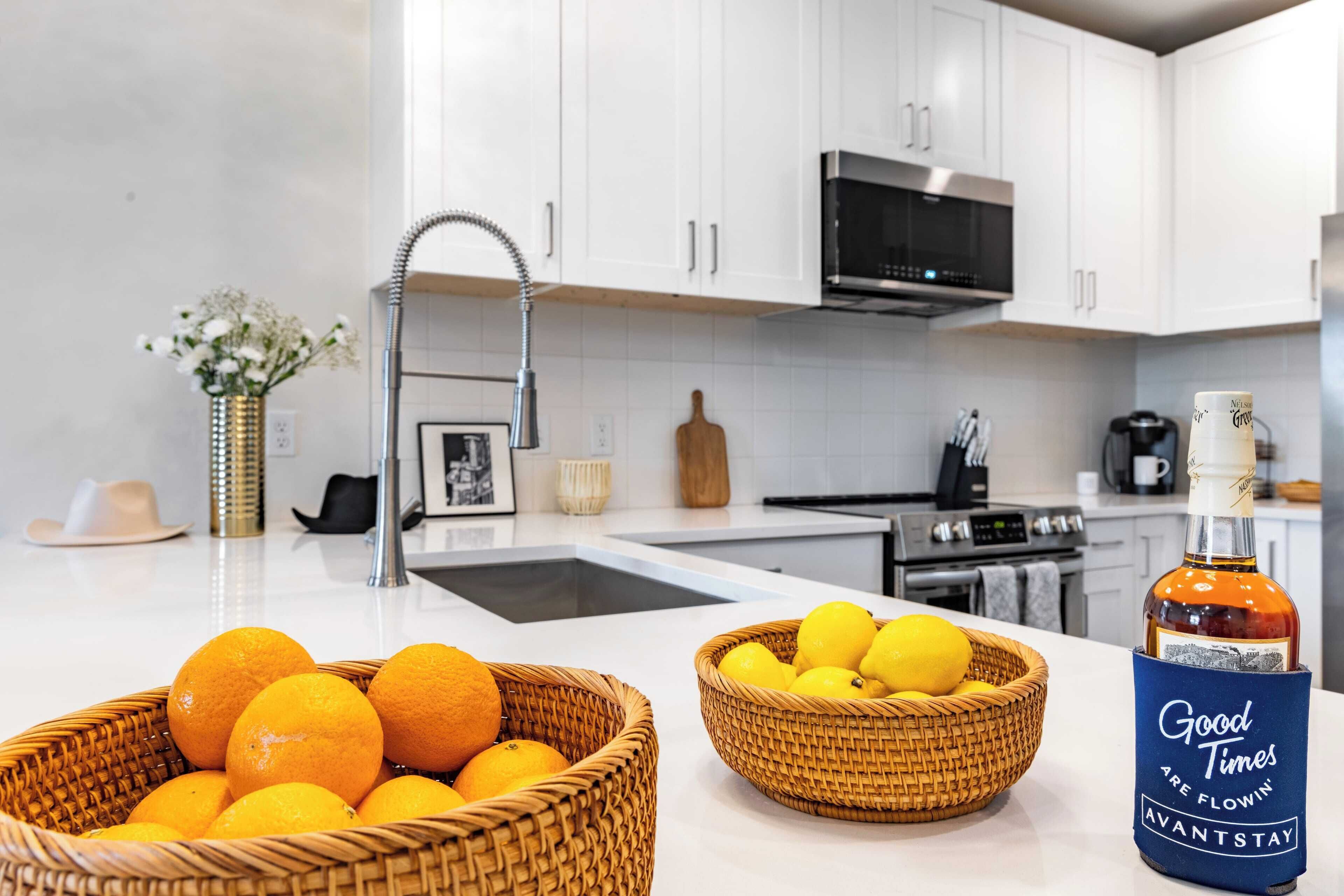 A modern kitchen with white cabinets, a stainless steel sink, and a microwave. There are two wicker baskets on the counter, one with oranges and the other with lemons. A bottle of liquor is also visible.