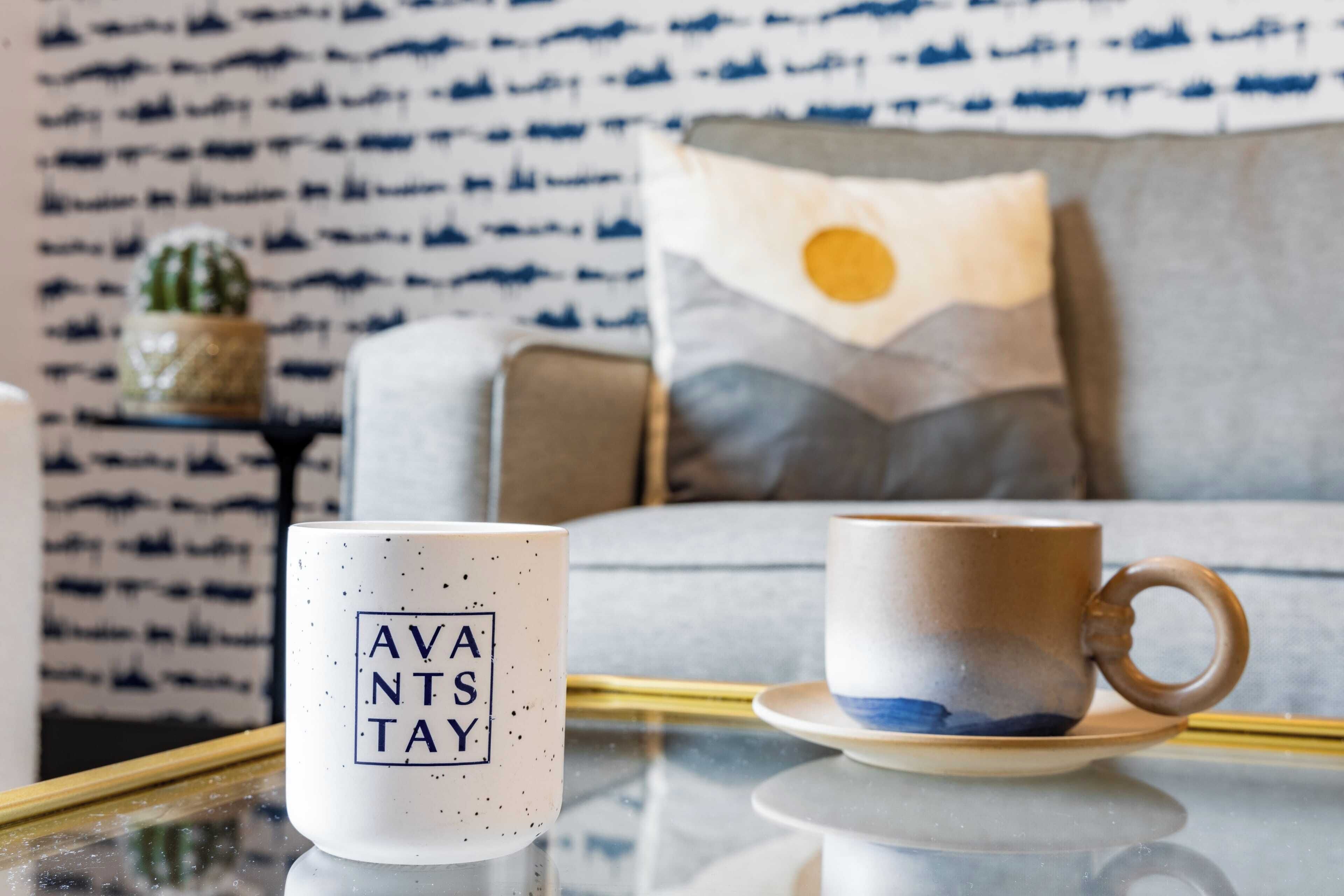 Een witte mok met 'AVANTS TAY', een bruine mok op een schoteltje, een kussen met een zon ontwerp, en een behang met patroon op de achtergrond.