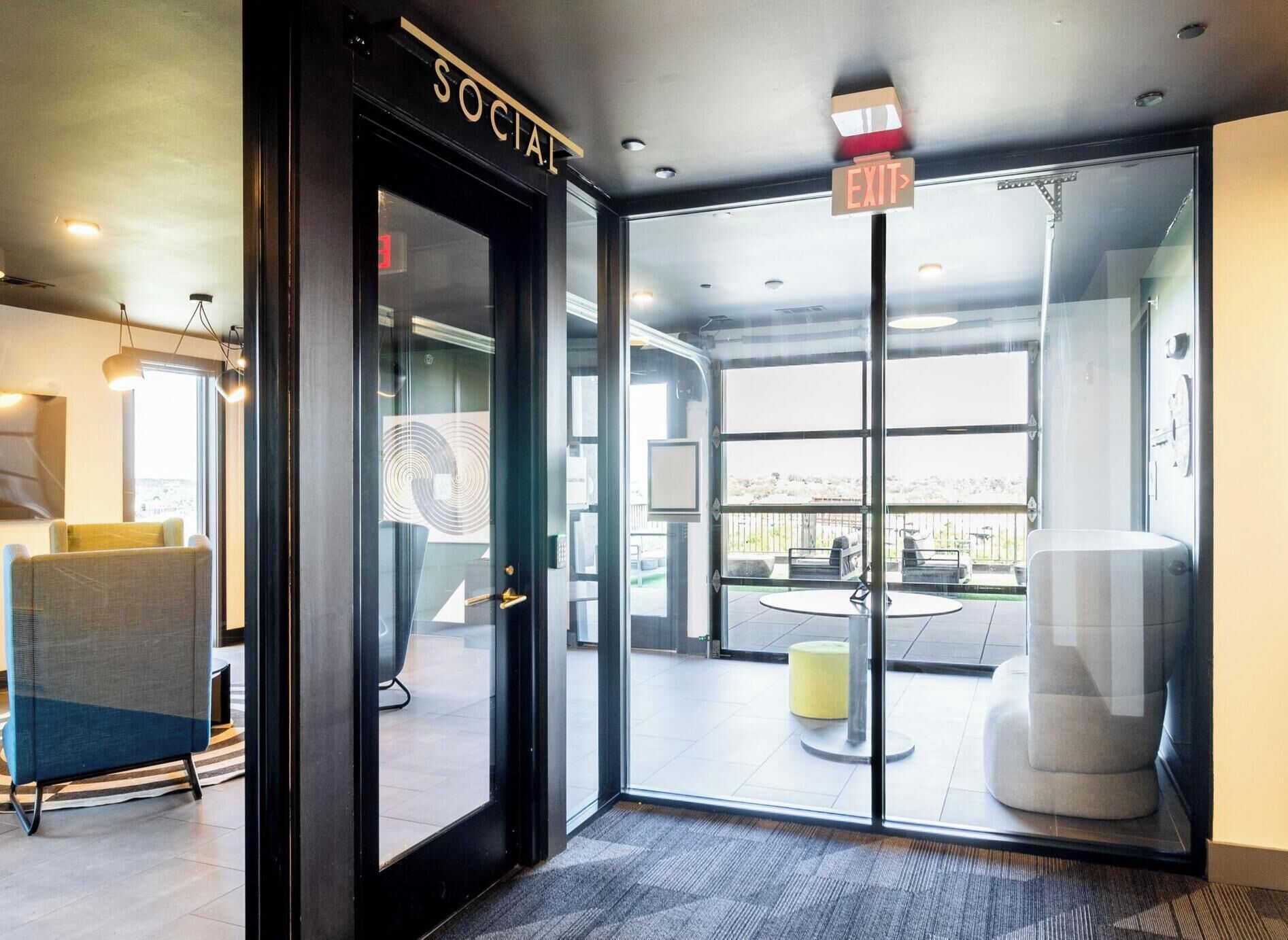 A modern building entrance with a glass door labeled 'SOCIAL,' an exit sign, and a view of an outdoor seating area.