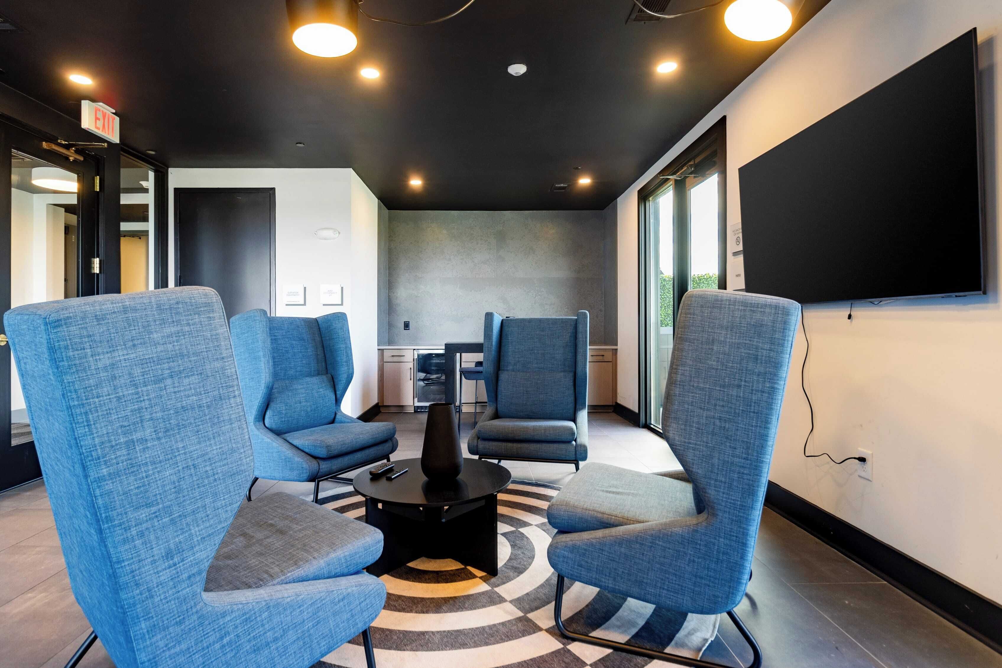 A modern meeting room with four blue chairs around a central table, a large blackboard on the wall, and a kitchen area in the background.
