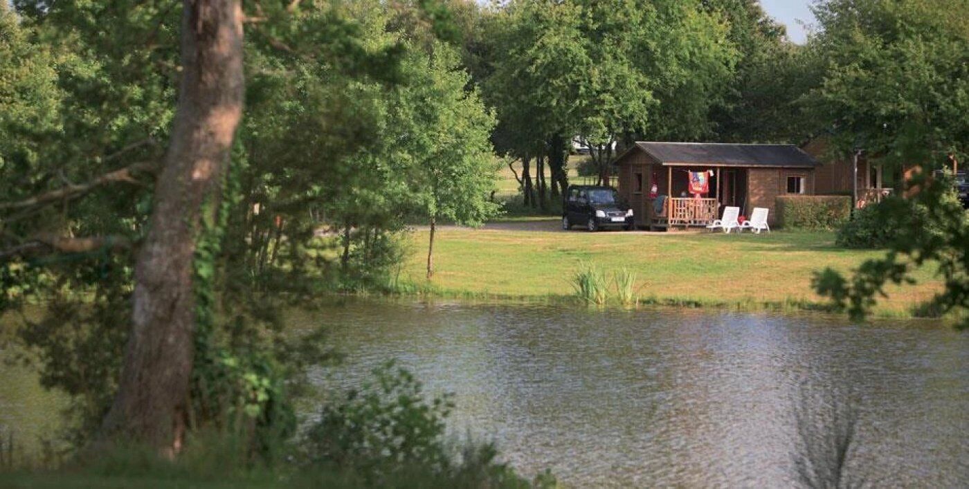 A cabin with a porch, a car parked nearby, and a pond in the foreground.