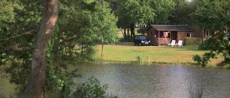 A cabin with a porch, a car parked nearby, and a pond in the foreground.