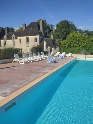 Una piscina con sillones de descanso, un edificio con techo de tejas y follaje al fondo.