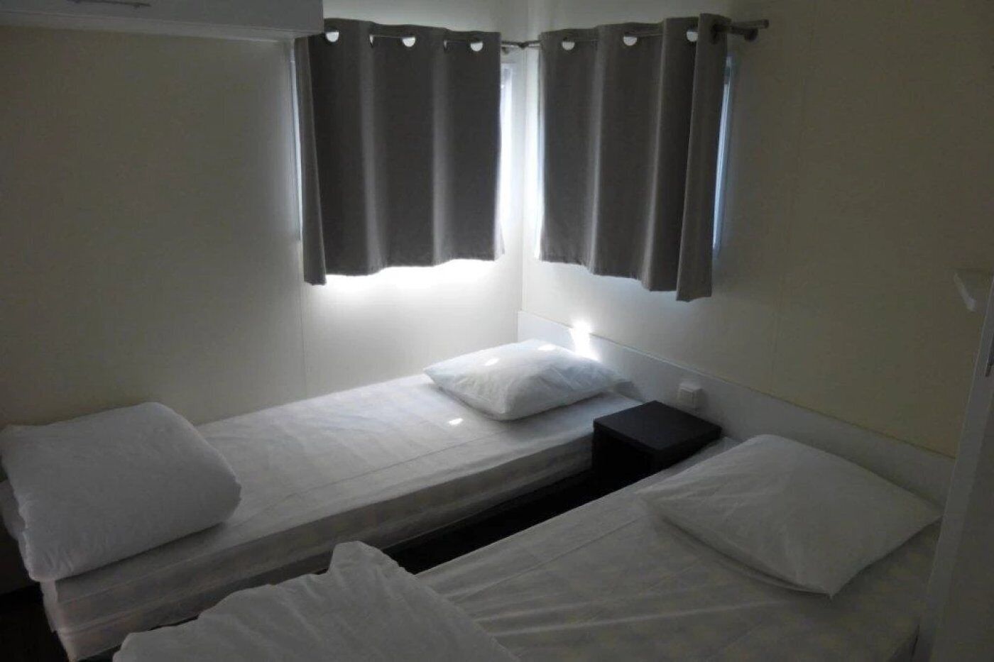 A small, single-room with two beds, a nightstand, and a window with curtains.
