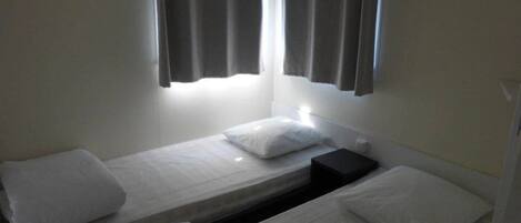 A small, single-room with two beds, a nightstand, and a window with curtains.