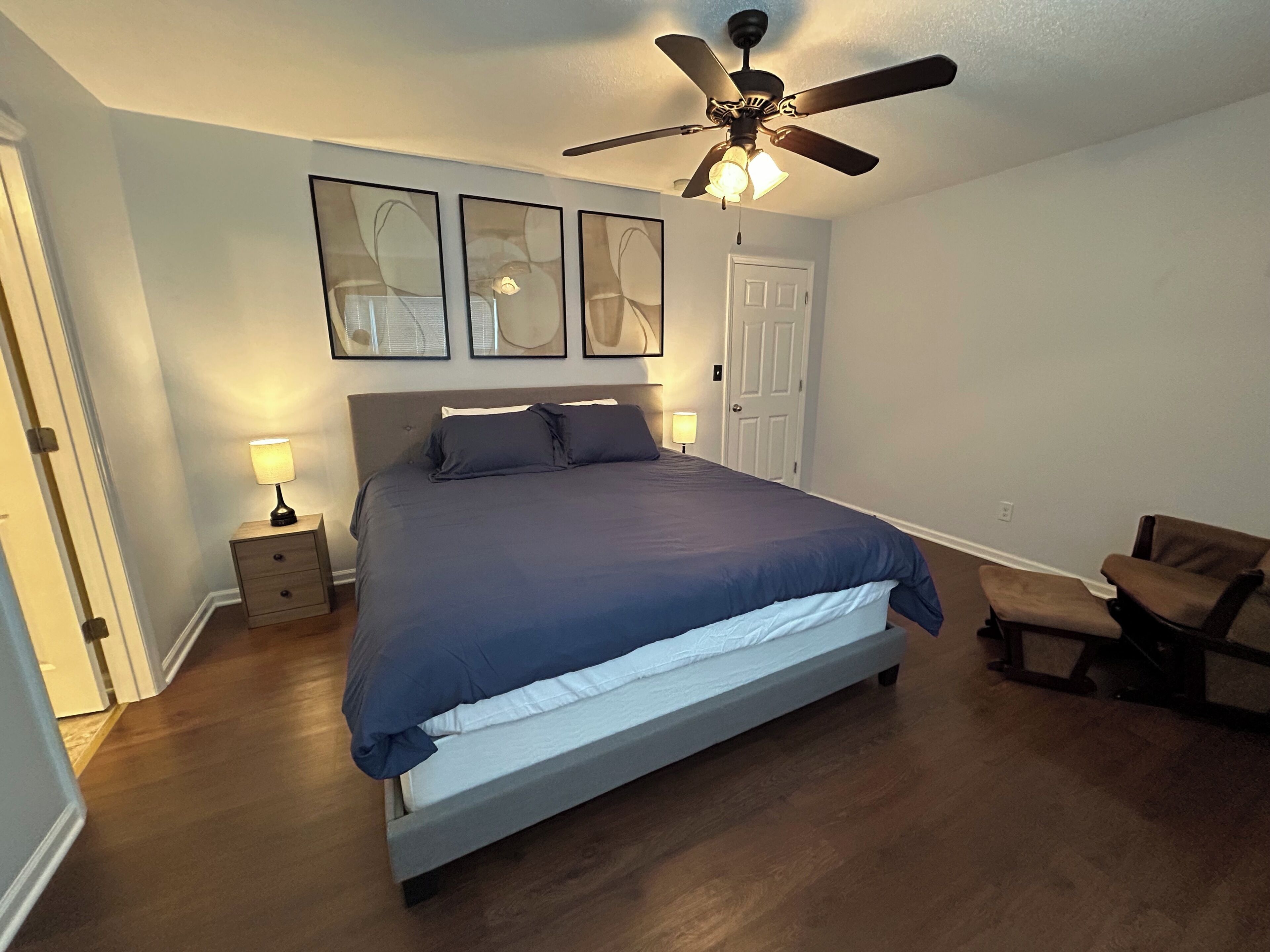 A bedroom with a bed, a ceiling fan, bedside lamps, a door, and a chair.