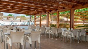 An outdoor dining area with tables and chairs, wooden ceiling beams, and a view of greenery.