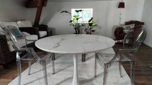 A round white table with a marble top surrounded by four clear chairs, set on a white rug in a room with wooden flooring and a window in the background. - Suite de Charme en Mezzanine (Remiremont)