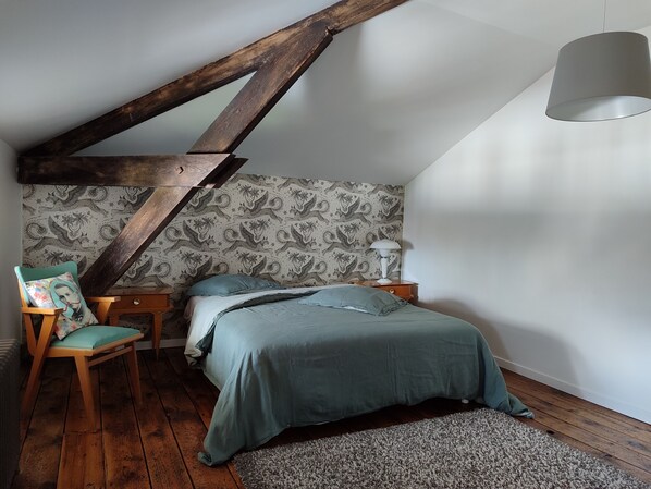 Ein Schlafzimmer mit schräg versiegelter Decke, einem Bett mit grüner Bettdecke, einem Sessel mit Blumenkissen, einem Nachttisch mit Lampe und einer Wand mit gemustertem Tapetenmuster.