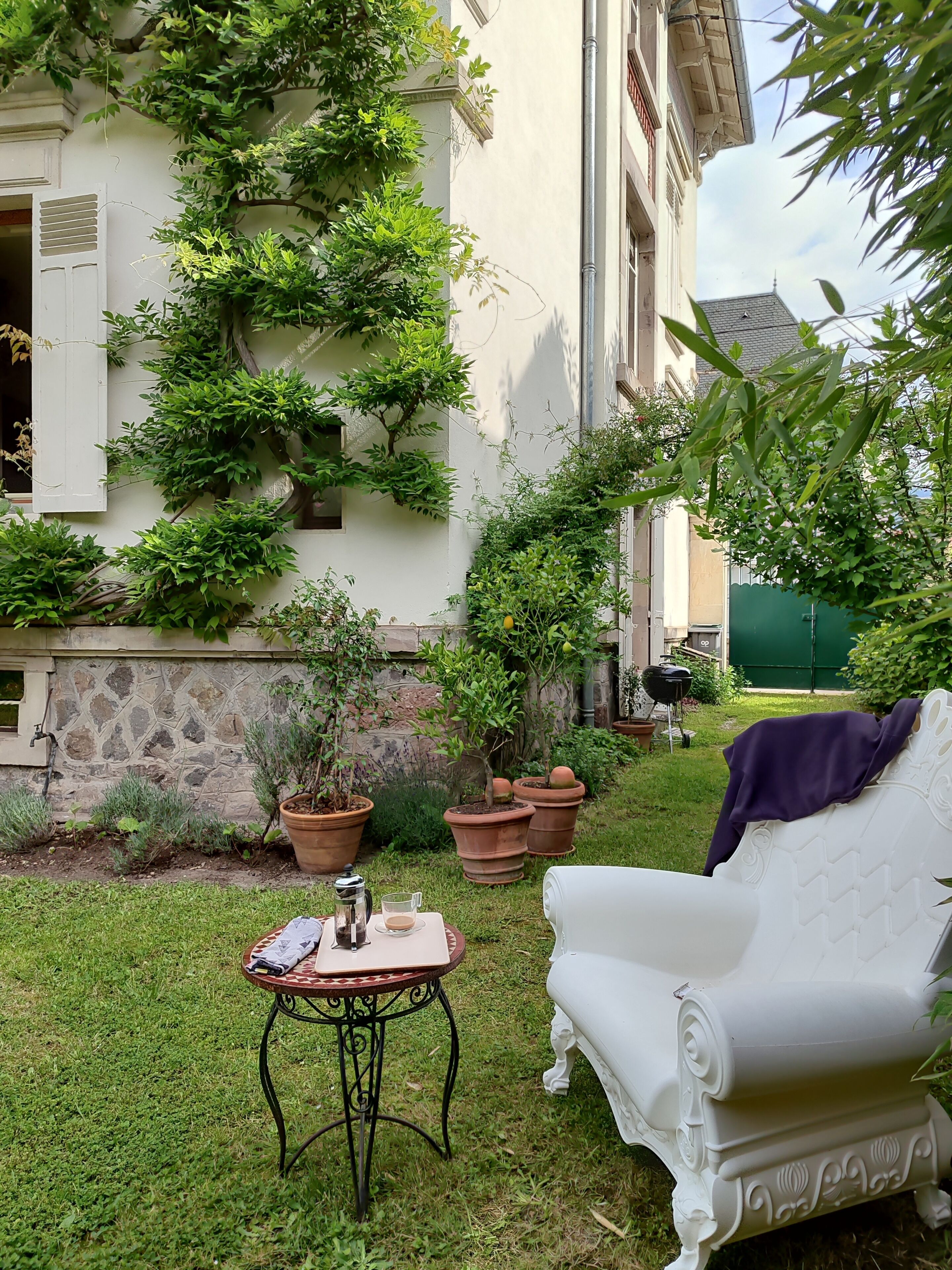 A white outdoor seating area with a table and a chair, surrounded by potted plants and a building with climbing vegetation.