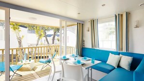 A room with a blue sofa, a dining table with chairs, and a view of the outdoors. - ZEcamping 3 étoiles piscine animations locatif TV LV clim 33m2 4 pers (Nailloux)