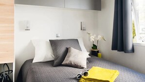 A neatly arranged bedroom with a bed, pillows, a small table with a vase of flowers, and a window with curtains. - ZEcamping 3 étoiles piscine animations locatif TV LV clim 33m2 4 pers (Nailloux)