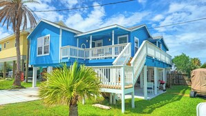 A two-story house with a balcony, a porch, and a small garden area. - Port A Cottage by Avantstay Private Patio + Deck Designed for Groups (Port Aransas)