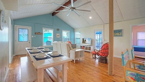 A spacious living area with a dining table set for four, a high ceiling with a fan, and a view of the outdoors through large windows. - Port A Cottage by Avantstay Private Patio + Deck Designed for Groups (Port Aransas)