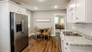 Una cocina moderna con armarios blancos, un refrigerador de acero inoxidable, un horno empotrado y un comedor con sillas y una mesa de madera.