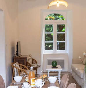 Een eethoek met een tafel gedekt voor twee personen, een fles drank en uitzicht op groen buiten het raam.