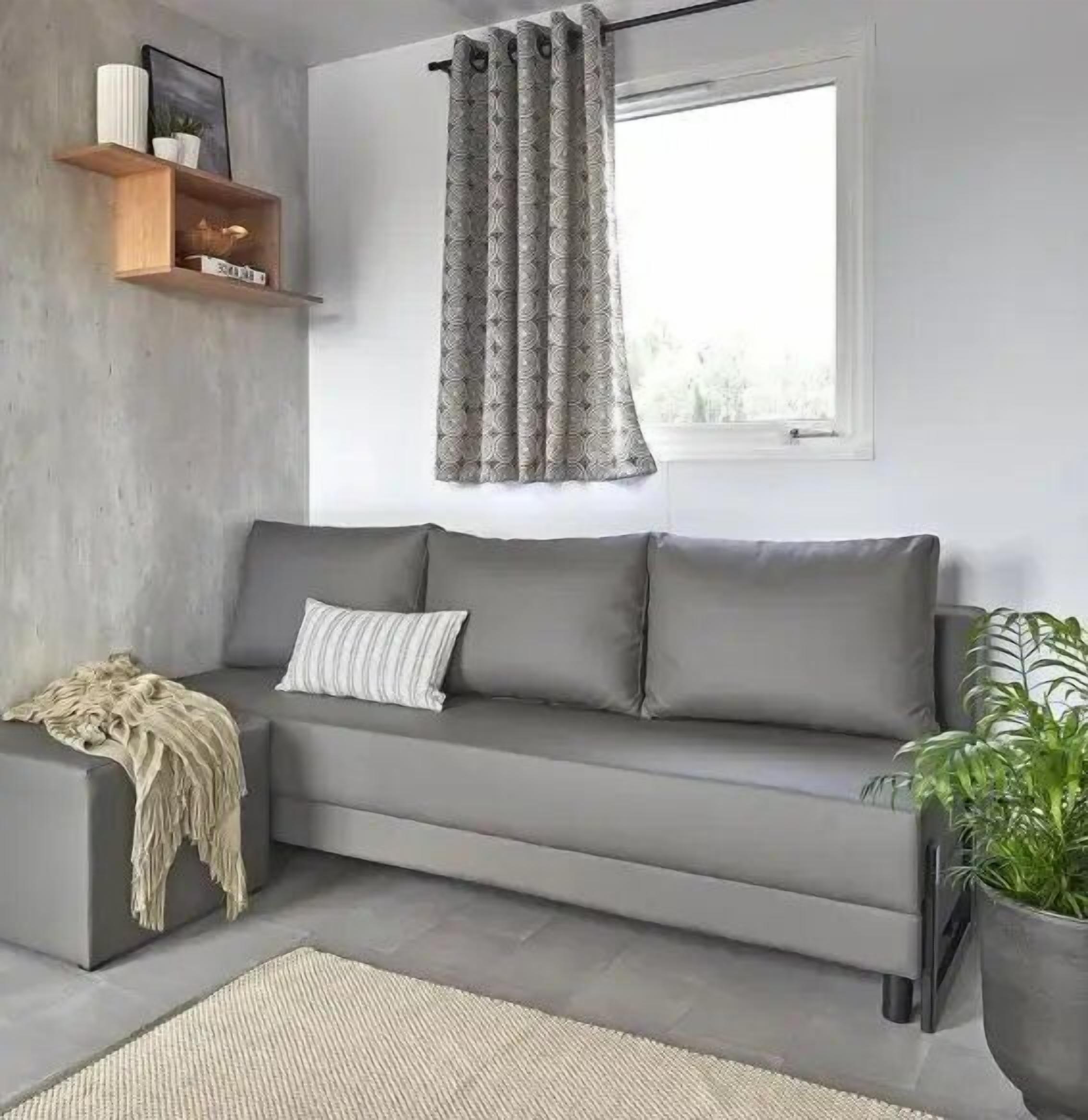 Una sala de estar moderna con un sofá gris, una manta, una alfombra pequeña, una planta en maceta y un ventanal con cortinas estampadas.