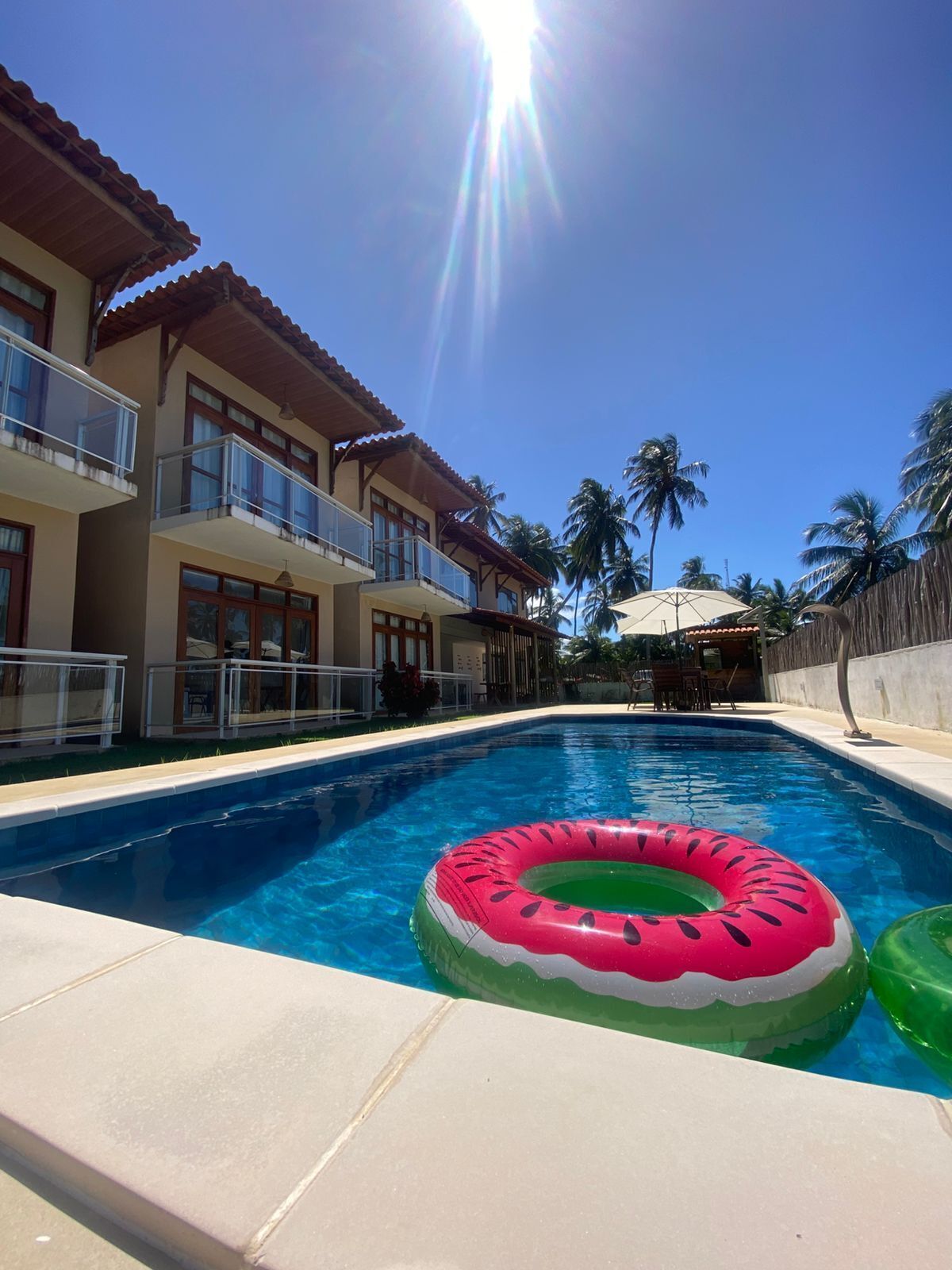 Una piscina con un flotador inflable rosa y verde, rodeada por un edificio con balcones y palmeras.