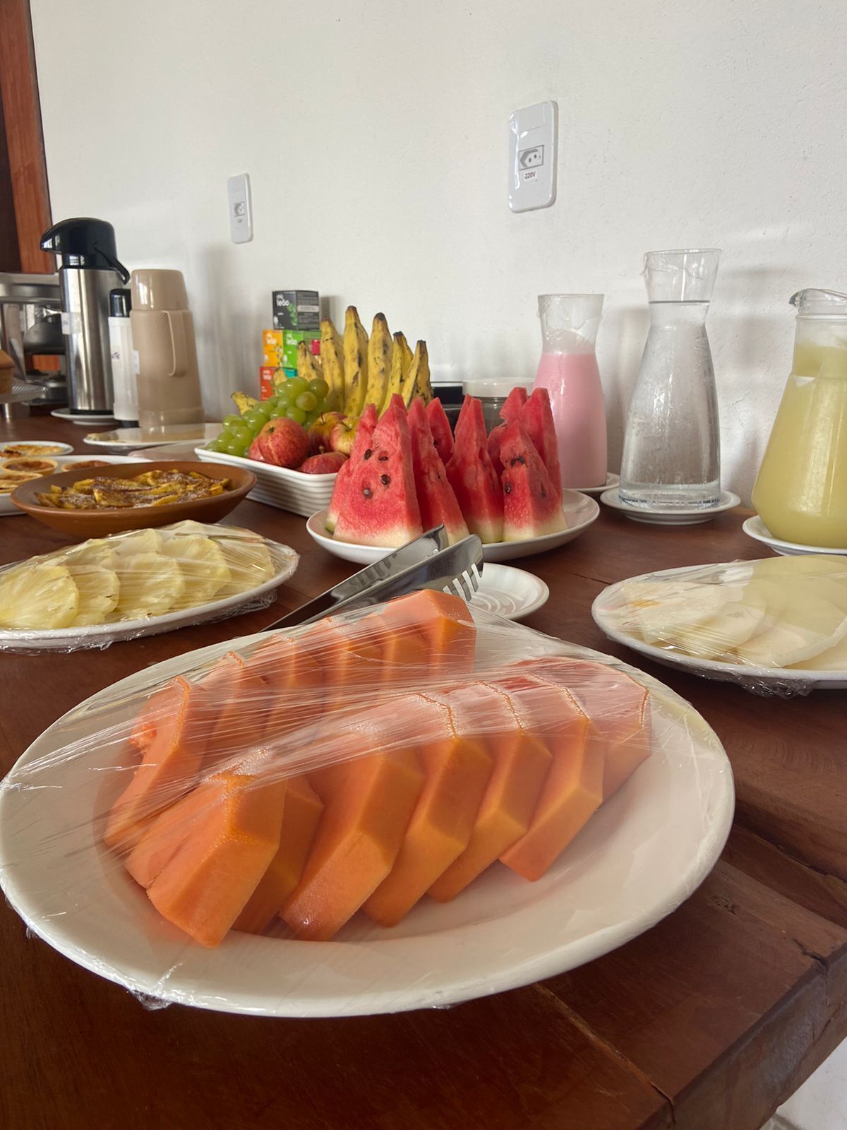 Colazione a buffet con diversi tipi di frutta, tra cui anguria, banane e uva, e bevande come succo di frutta e caffè.