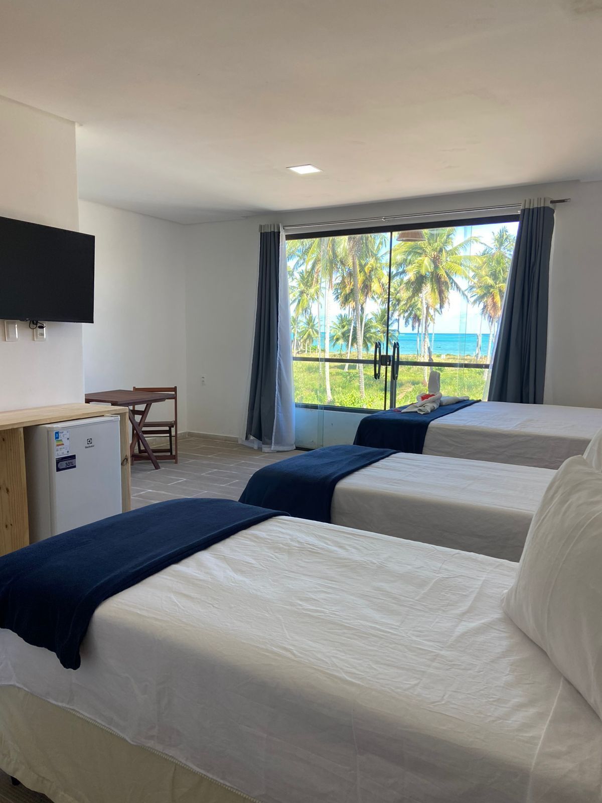 Habitación de hotel con dos camas, un escritorio, un televisor y una ventana con vista a palmeras y al océano.