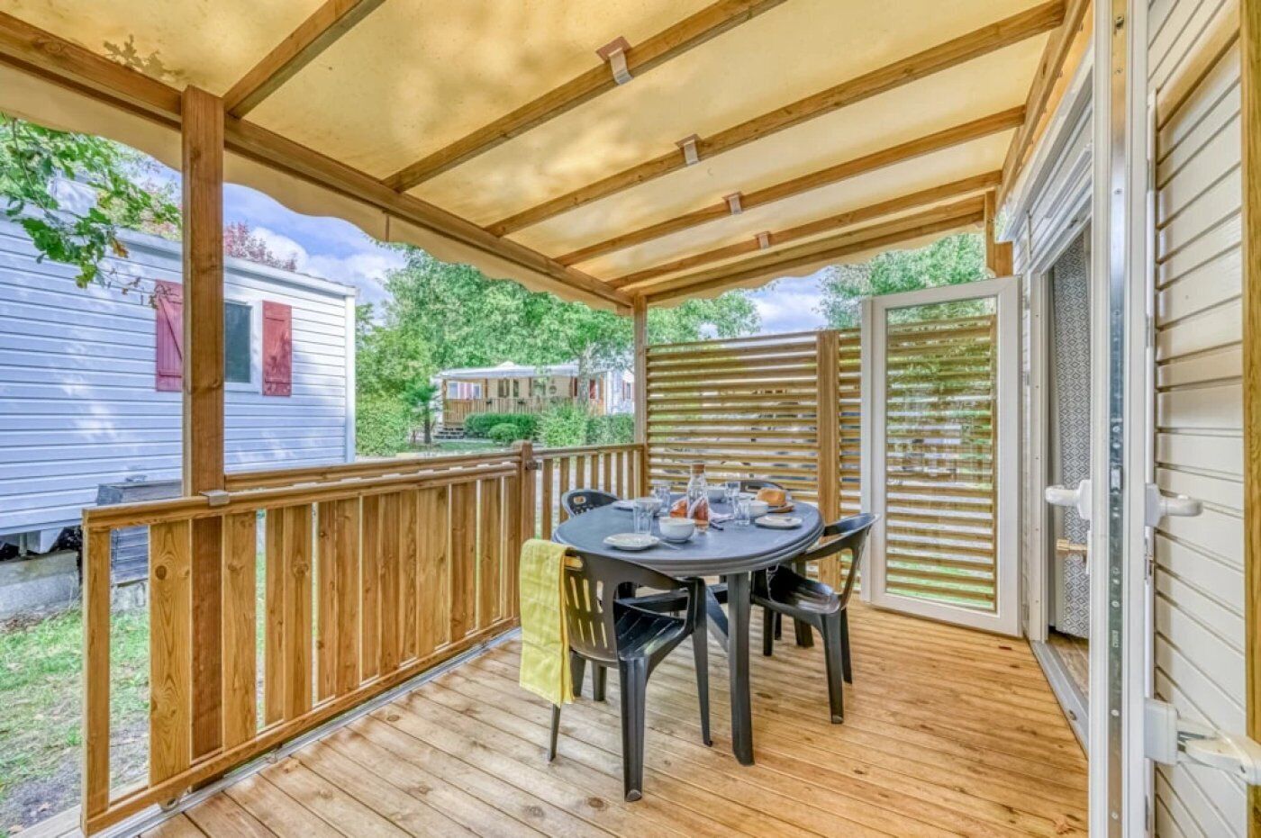 Une terrasse extérieure couverte, dotée d’une table et de chaises, de rampes en bois et offrant une vue sur les maisons voisines.