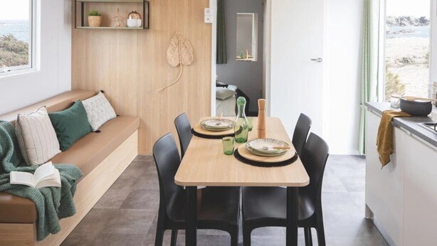 Ein modernes Interieur mit einer Holzwand, einem Esstisch mit Tellern und Gläsern, schwarzen Stühlen und einem grünen Sofa mit Kissen.
