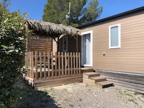 Una casa móvil de un nivel con una terraza de madera, una estructura de techo de paja y una puerta blanca.