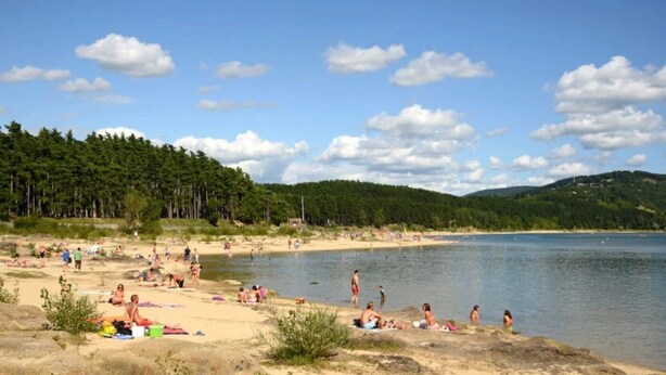 A beach with people sunbathing and swimming, surrounded by trees and hills.
