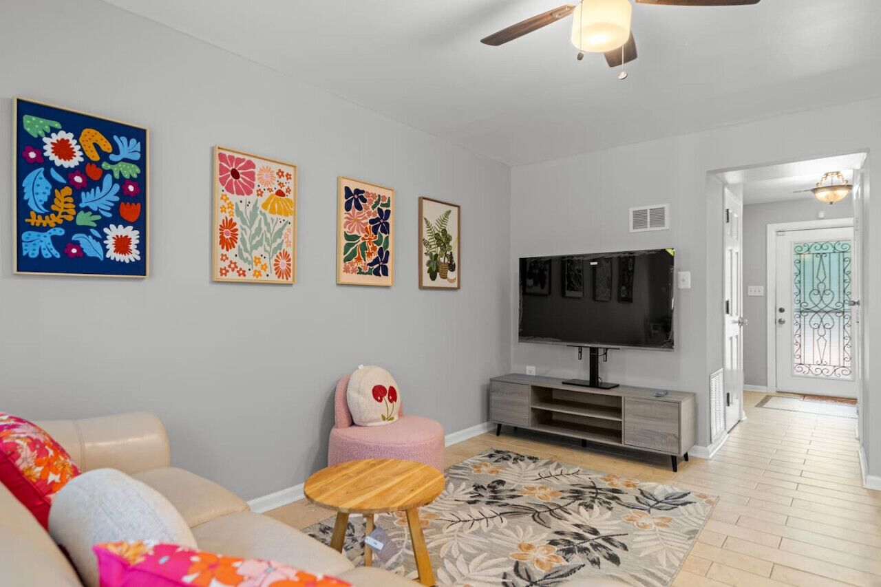 Una sala de estar con un sofá color beige, un reposapiés rosa, una mesa de centro de madera, un televisor de pantalla plana y decoración floral en la pared.
