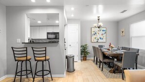A modern kitchen with white cabinets, a black microwave, and a stainless steel sink. There are two bar stools and a dining table with chairs. A chandelier hangs above the table, and a potted plant is on the floor.
