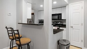 A compact kitchen with white cabinets, a microwave, and a stove. There is a trash can and a chair in the corner.